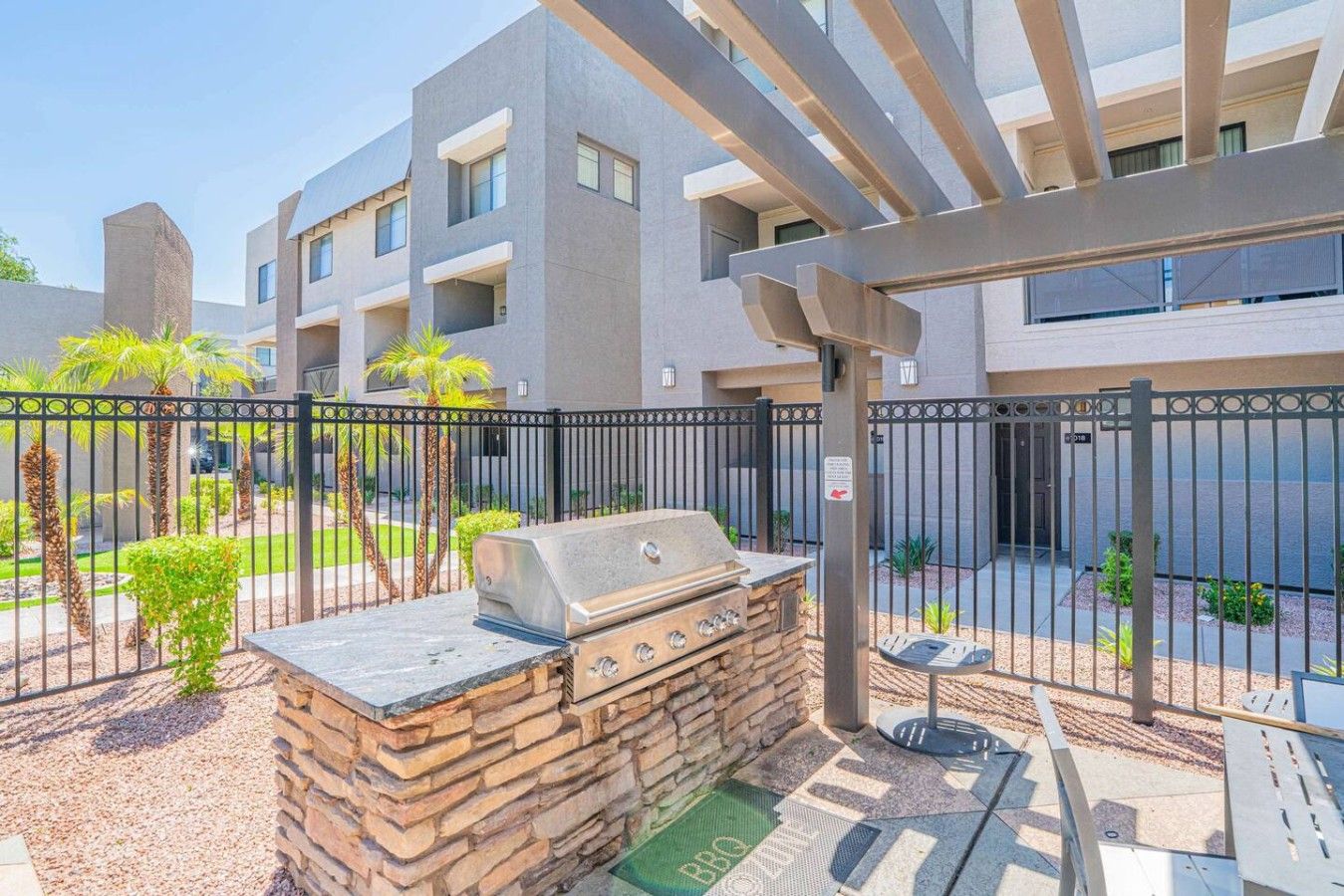 A barbecue area with a stone grill, a pergola, and a gated outdoor space with a building in the background.