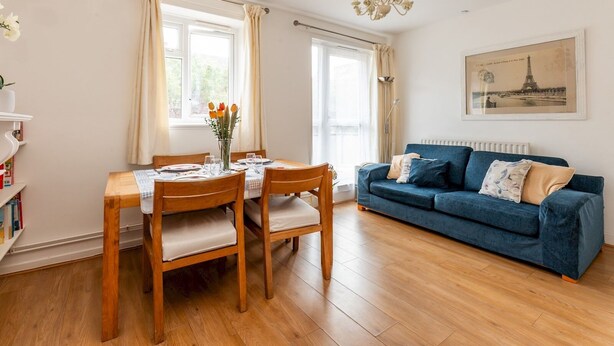 Un comedor con mesa y sillas de madera, un florero con flores y un sofá azul en una habitación luminosa con ventanas grandes y una araña.
