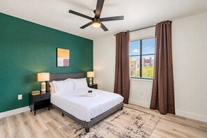 A hotel room with a bed, two bedside tables with lamps, a ceiling fan, a window with curtains, and a view of buildings outside.