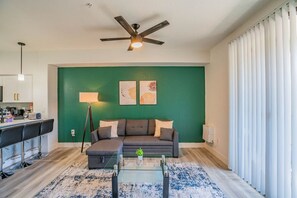 A living room with a green wall, a ceiling fan, a glass coffee table, a sofa, and a bar area with stools. - Lavish 2BD| Walk to Westgate+Pool+Gym+W/D (Glendale)