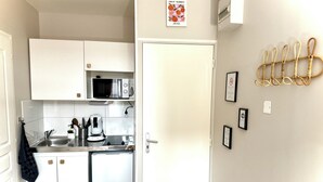 A compact kitchen with white cabinets, a microwave, a coffee maker, and a refrigerator. There are framed pictures on the wall and a fruit market poster above the door.