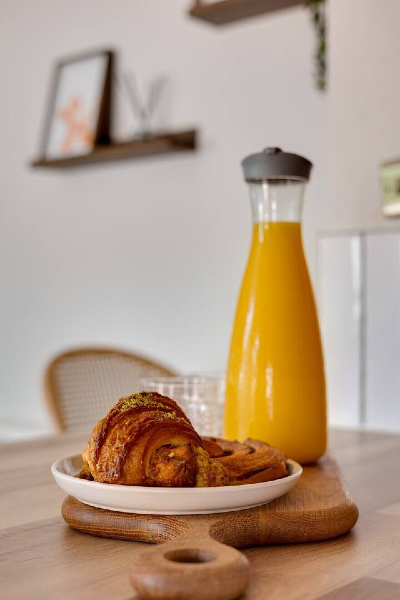A breakfast setting with a croissant on a wooden board, a glass of orange juice, and a coffee maker.