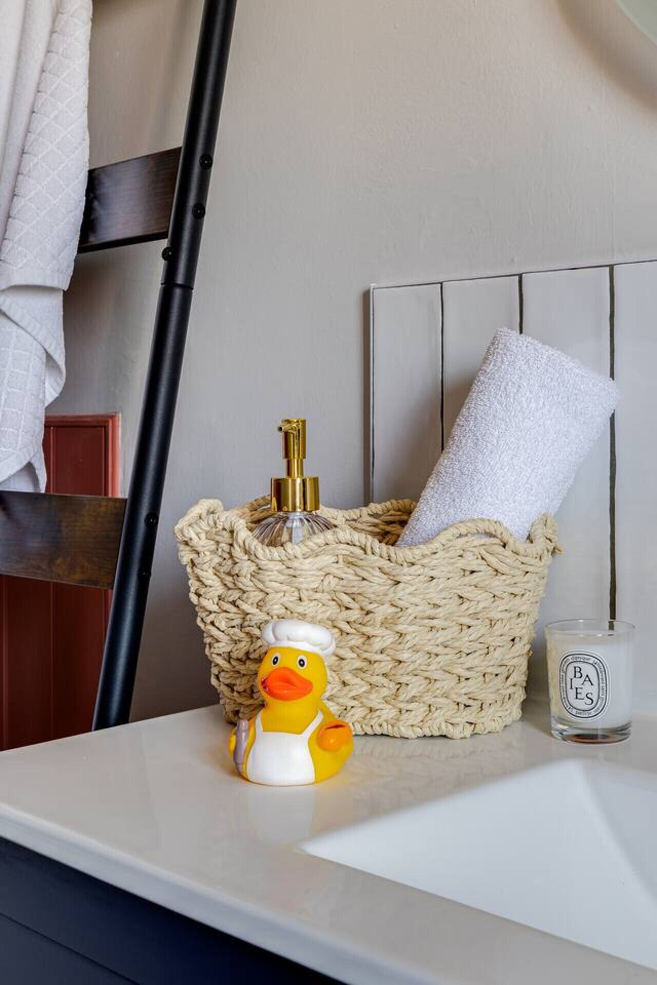 A bathroom with a woven basket containing a towel and a bottle, a duck figurine, and a glass of water on a white countertop.