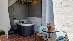 A hot tub with a rustic brick wall, a small table with a potted plant, and a chair with patterned cushions.