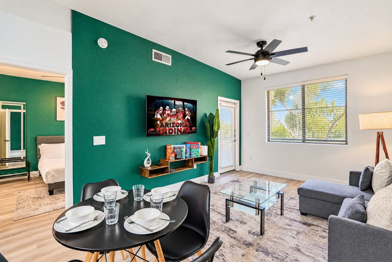 A living room with a green accent wall, a flat-screen TV, a dining area with a table set for two, and a gray sofa.