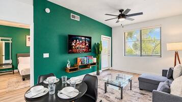 A living room with a green accent wall, a flat-screen TV, a dining area with a table set for two, and a gray sofa.