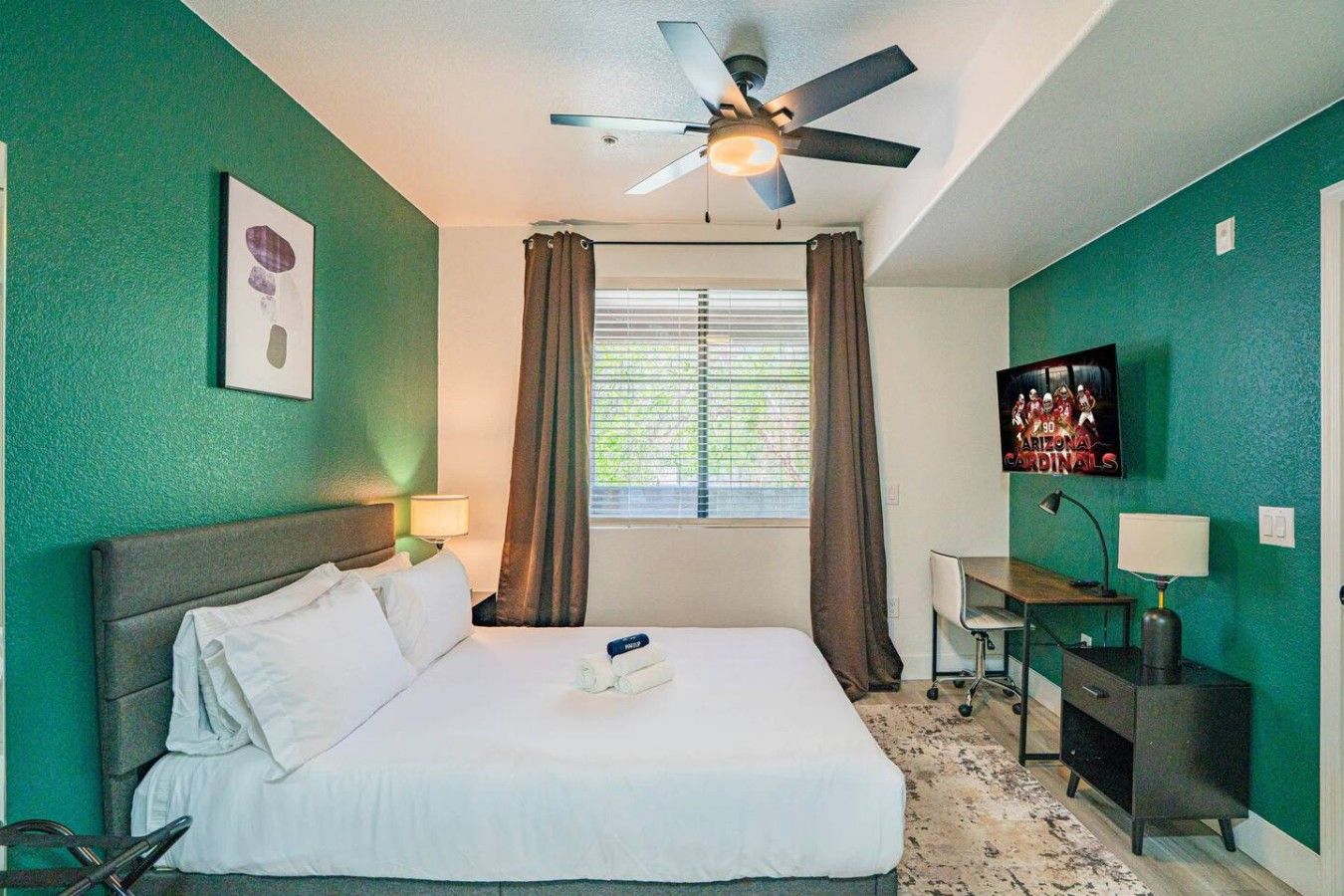 A hotel room with a bed, a desk, a ceiling fan, a window with curtains, and a television on the wall.
