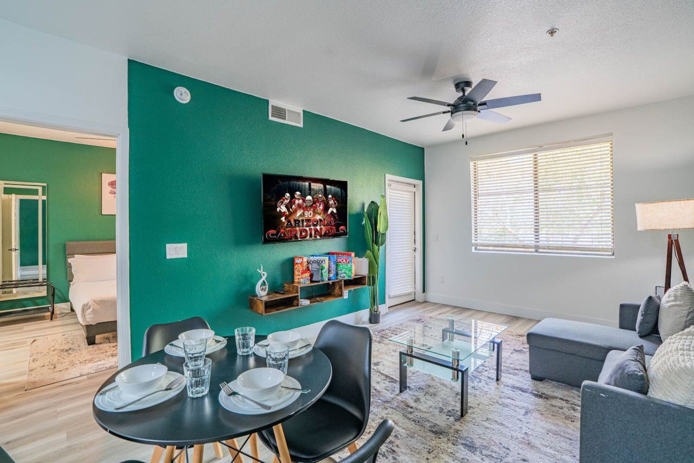 A living room with a teal accent wall, a ceiling fan, a television, and a dining area with a glass table and chairs.