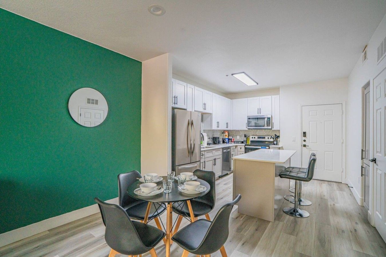 A modern kitchen with white cabinets, a stainless steel refrigerator, and a microwave. There is a dining area with a round table and black chairs.