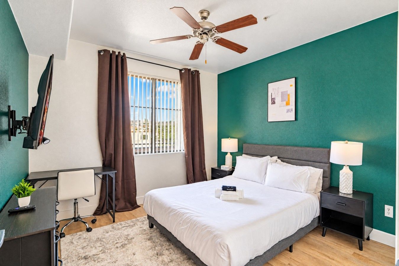A hotel room with a bed, two bedside tables with lamps, a desk with a chair, a ceiling fan, a window with curtains, and a wall-mounted TV.