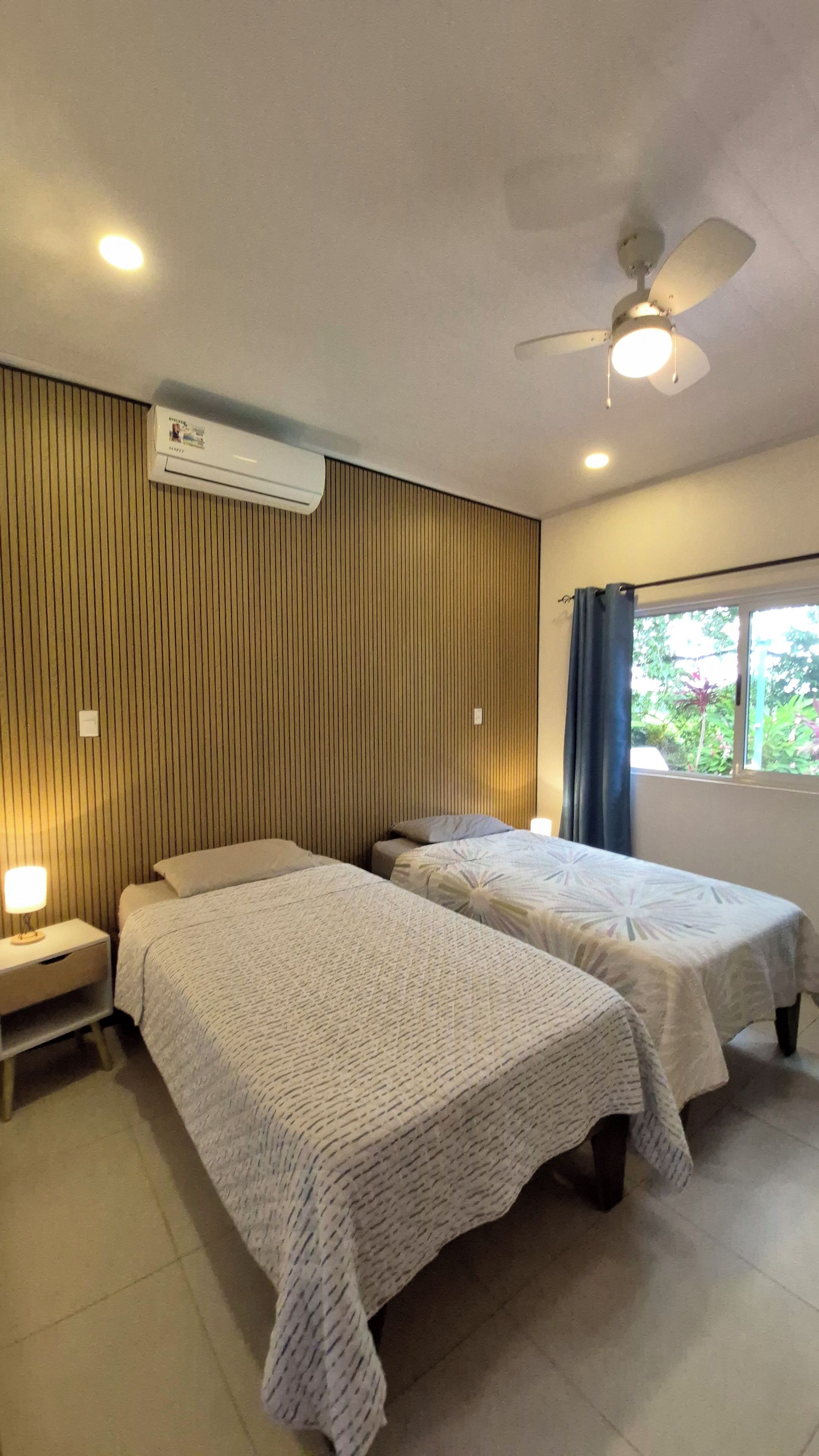 Une chambre d’hôtel avec deux lits simples, un ventilateur de plafond, un appareil de climatisation, une fenêtre avec des rideaux et une table de chevet avec une lampe.