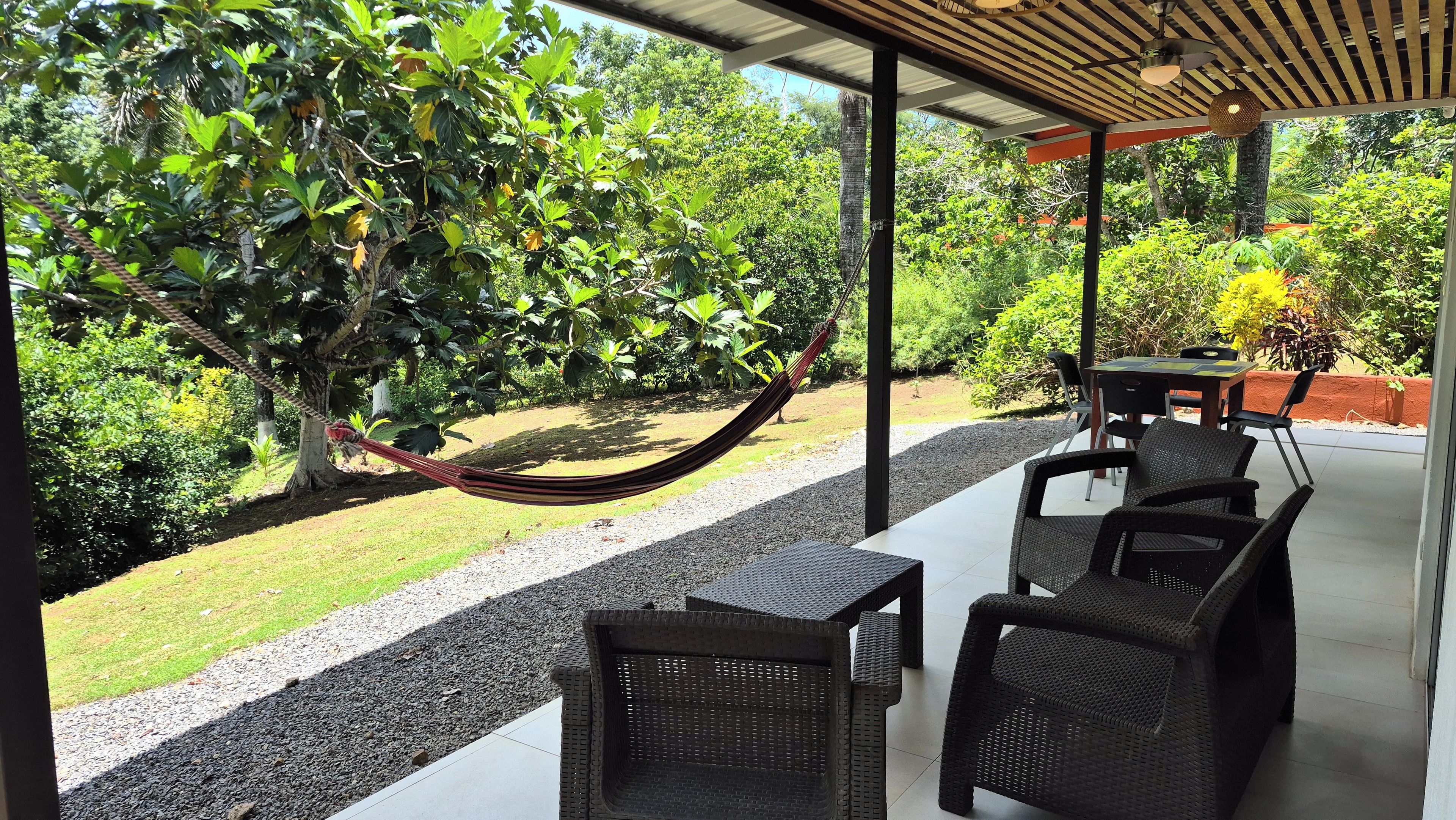 A covered outdoor seating area with a hammock, wicker furniture, and a view of a lush garden.