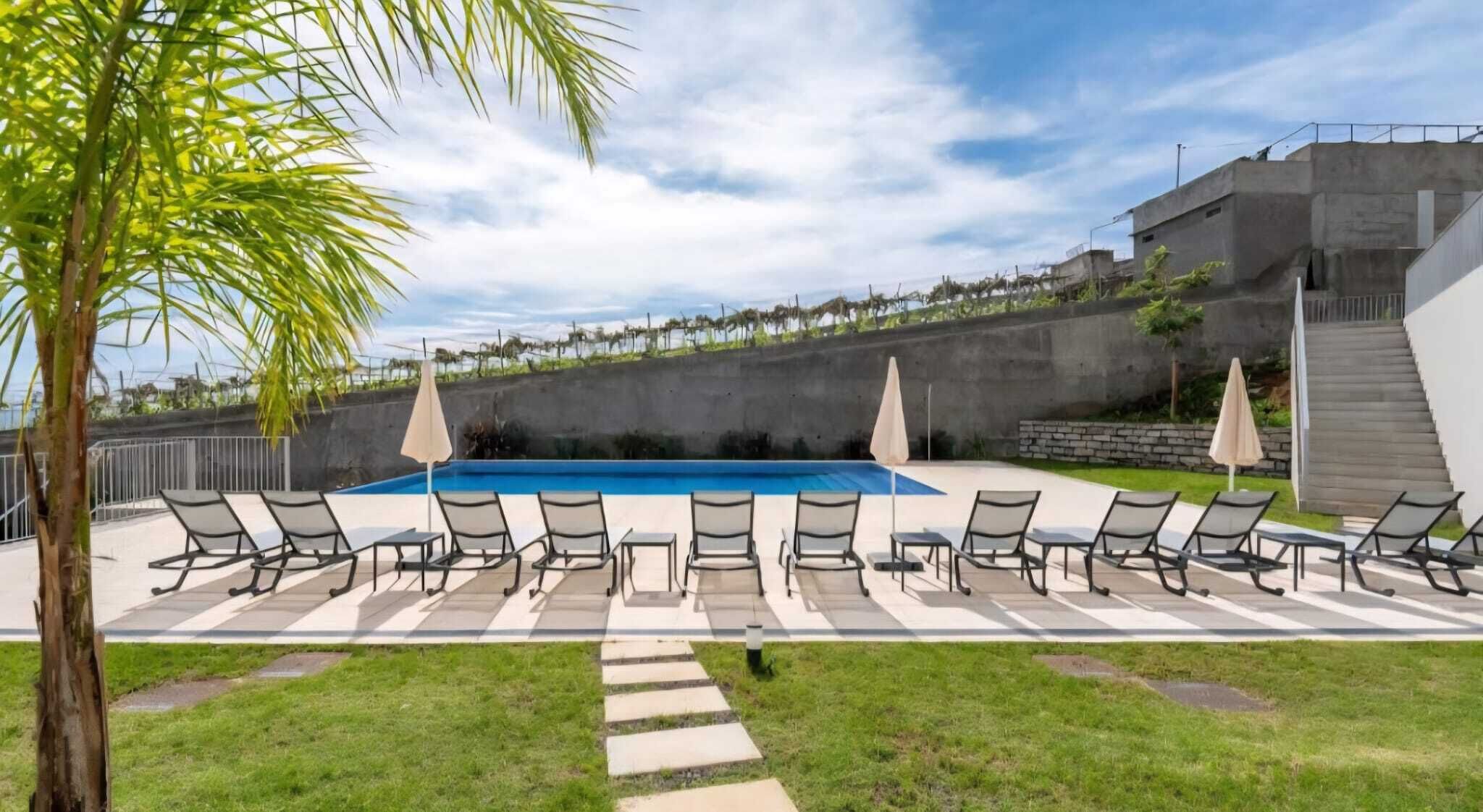 Una piscina con sillas de descanso y sombrillas, rodeada de césped y un muro de hormigón.