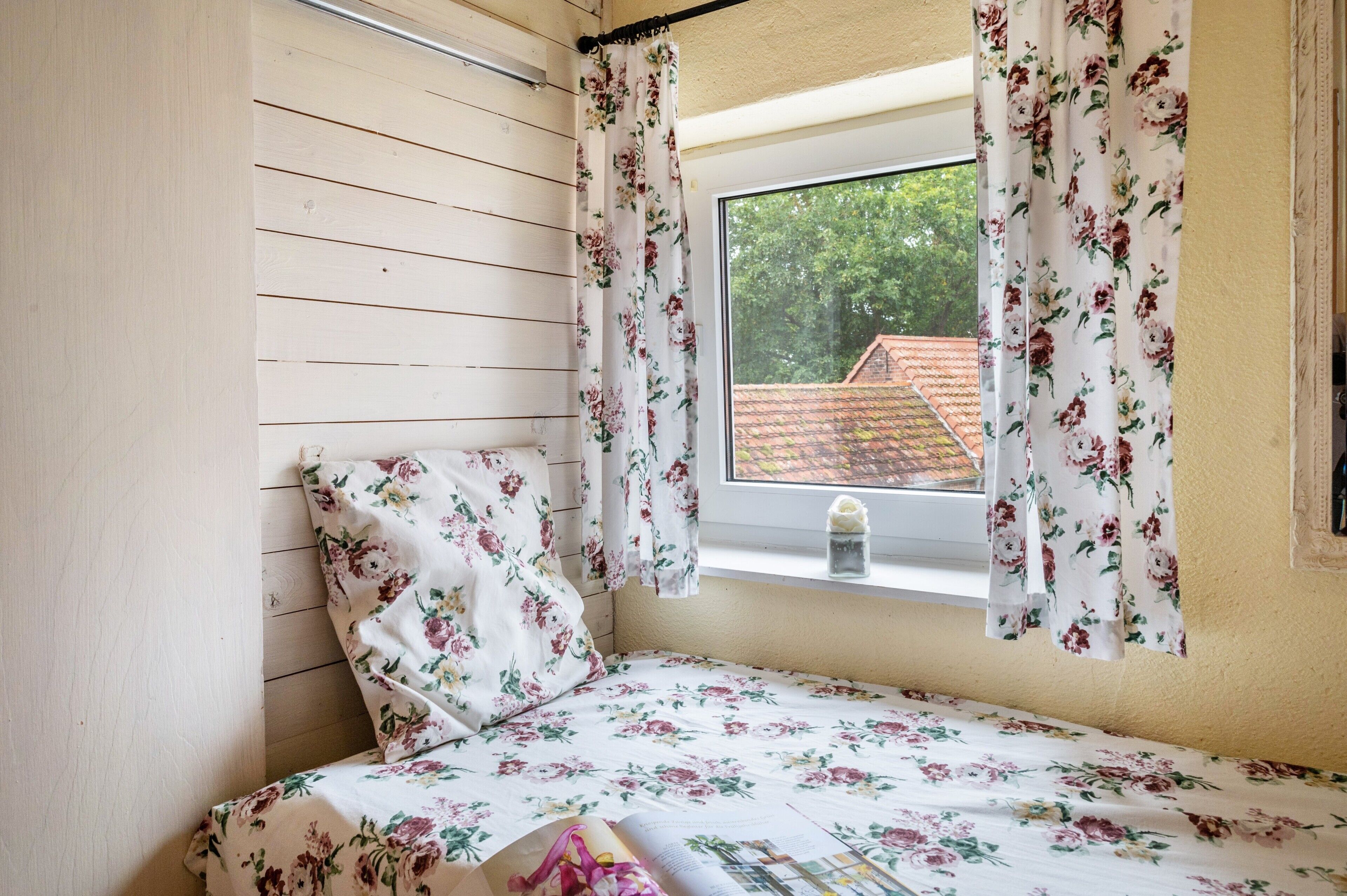 Une chambre avec un mur recouvert de lambris en bois, un lit avec une literie à motifs floraux, une fenêtre ornée de rideaux à fleurs et une vue sur un bâtiment aux toits rouges à l’extérieur.