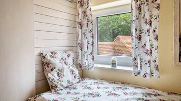 Une chambre avec un mur recouvert de lambris en bois, un lit avec une literie à motifs floraux, une fenêtre ornée de rideaux à fleurs et une vue sur un bâtiment aux toits rouges à l’extérieur.