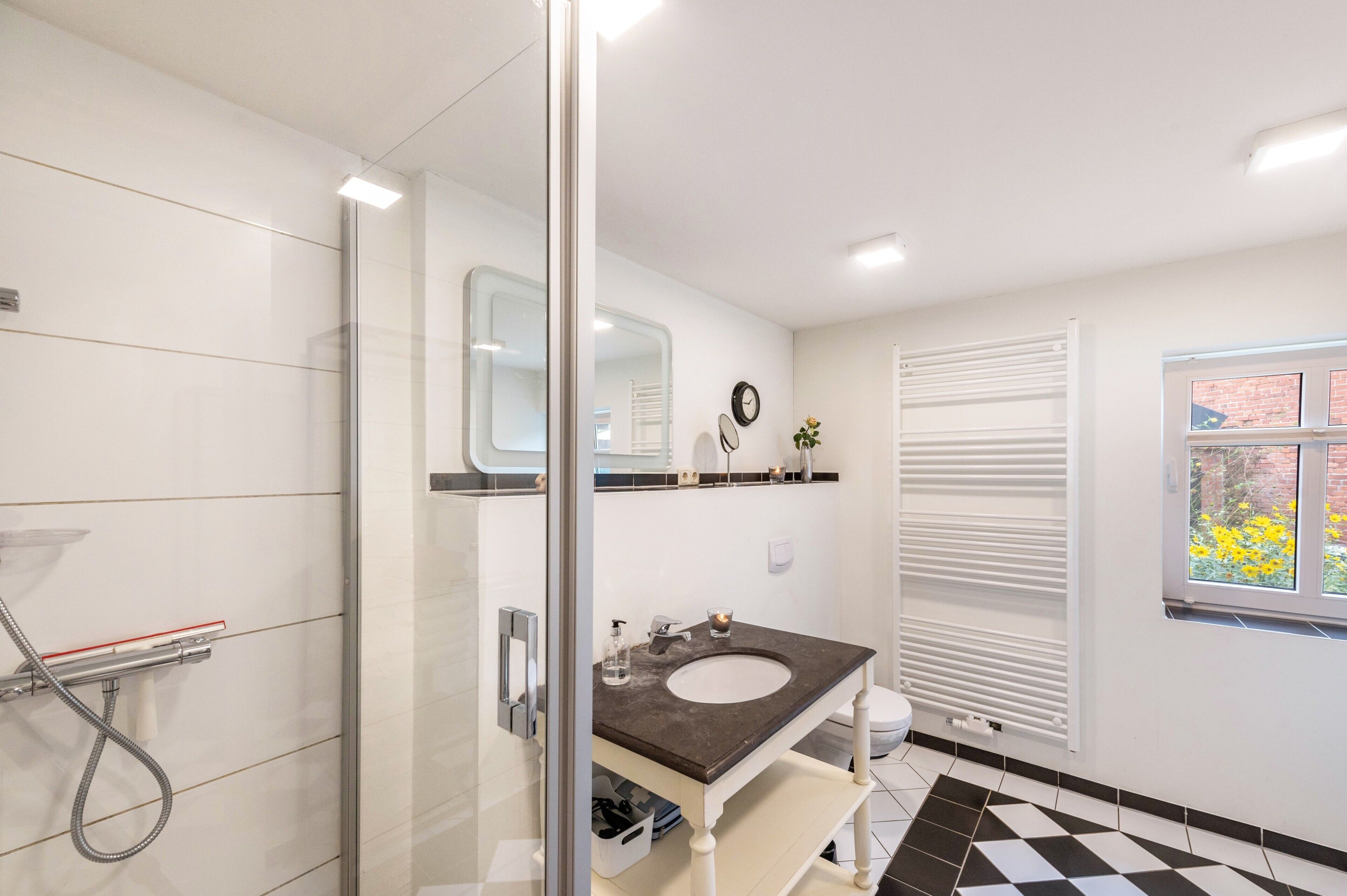Une salle de bain avec une douche à parois de verre, un lavabo doté d’un plan de travail foncé, un miroir, une horloge et une fenêtre donnant sur des fleurs.