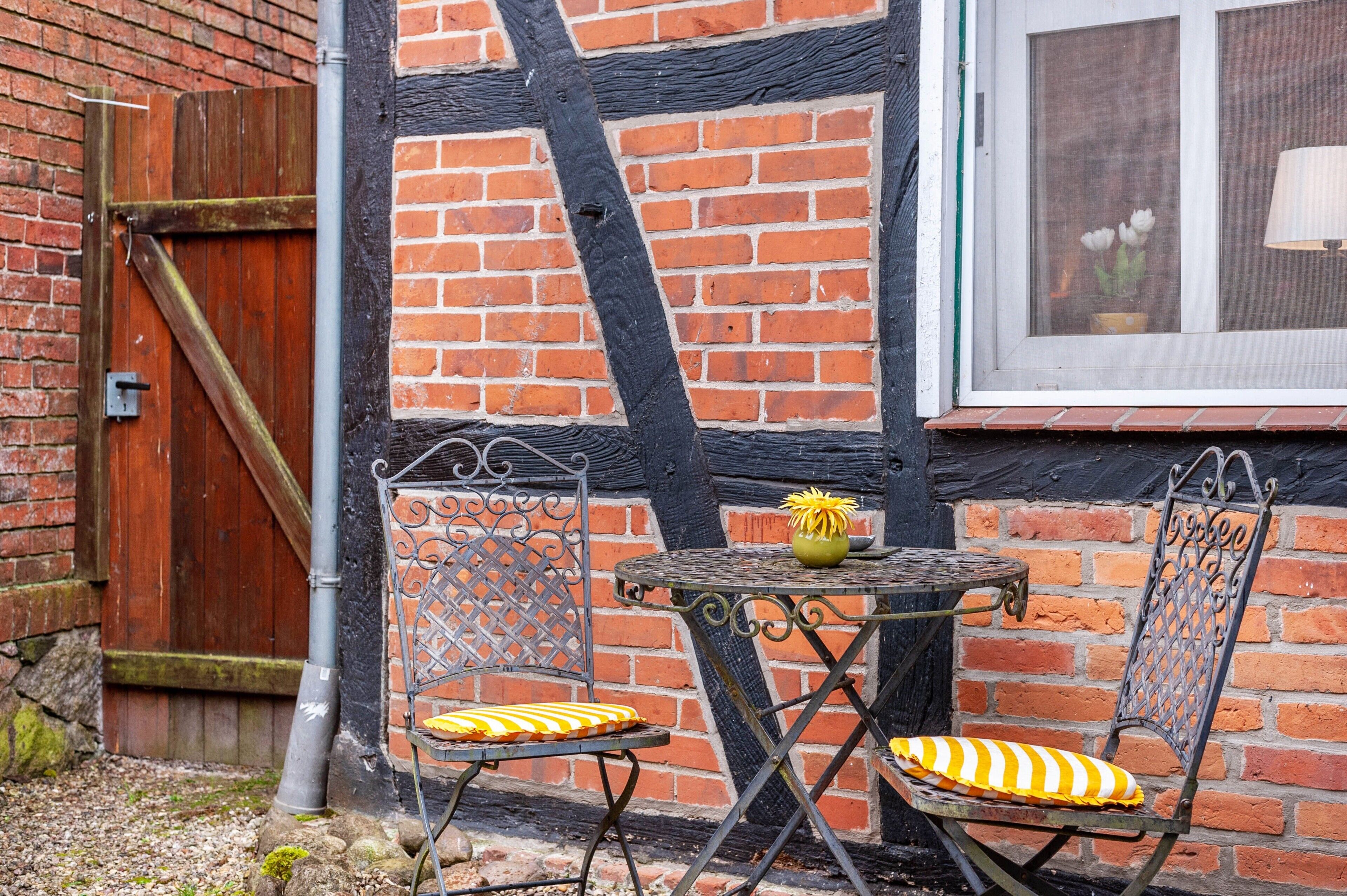 En rustik utomhusplats med sittmöbler, bestående av ett metallbord och stolar, en trädörr och en mur av tegelsten.