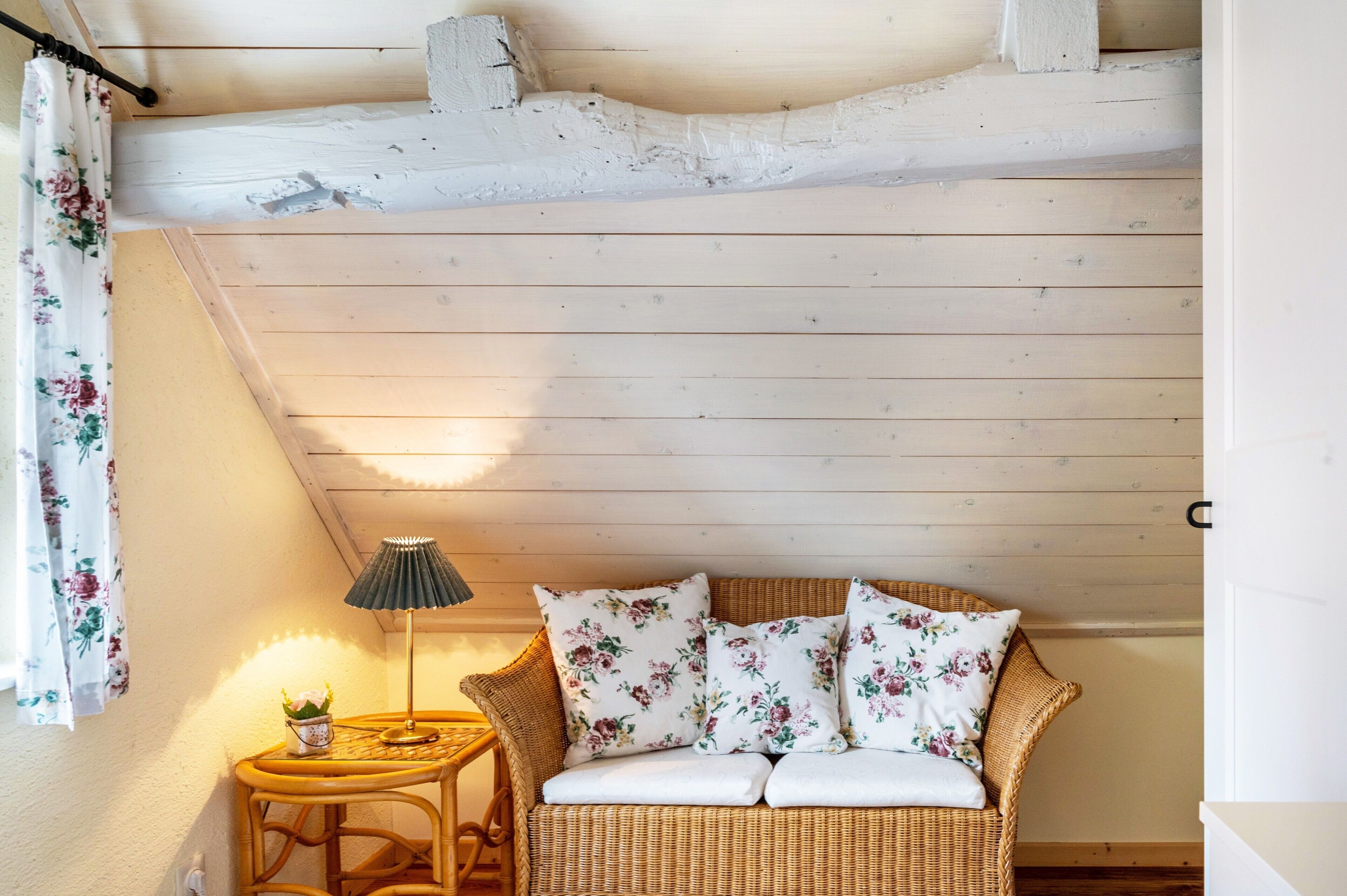 Ein Zimmer mit einem Korbssessel, floralen Kissen, einem kleinen Tisch mit Lampe und einem Vorhang.