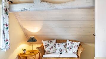 Ein Zimmer mit einem Korbssessel, floralen Kissen, einem kleinen Tisch mit Lampe und einem Vorhang.