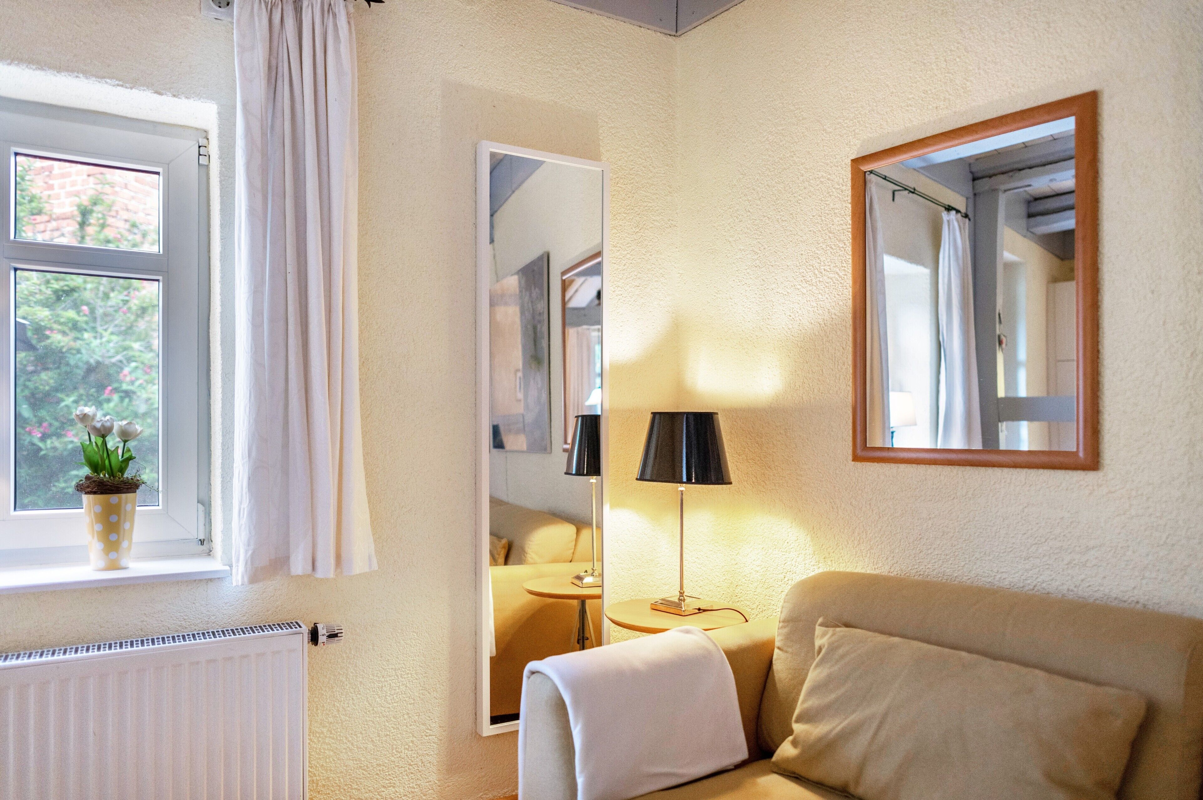 Una habitación con ventana, un radiador, una planta en maceta, un sofá, una lámpara y un espejo.