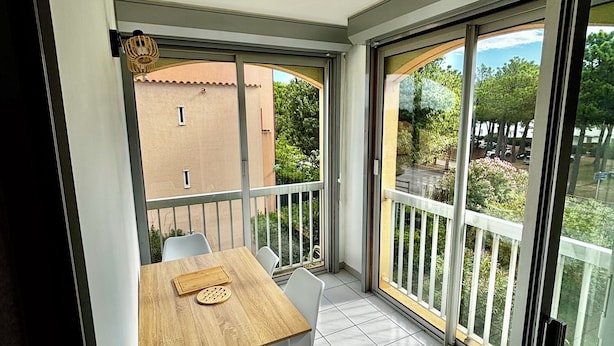 Un balcón con mesa y sillas de madera, vistas a zonas verdes y edificios, y una puerta de cristal.