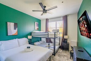 Een hotelkamer met een stapelbed, een eenpersoonsbed, een plafondventilator, een raam met gordijnen, een bureau met een lamp en een lijst aan de muur.