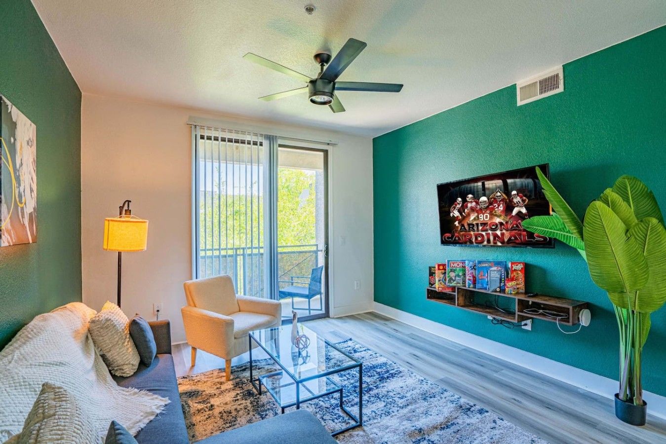 A living room with a sofa, armchair, glass coffee table, and a TV mounted on a teal wall.