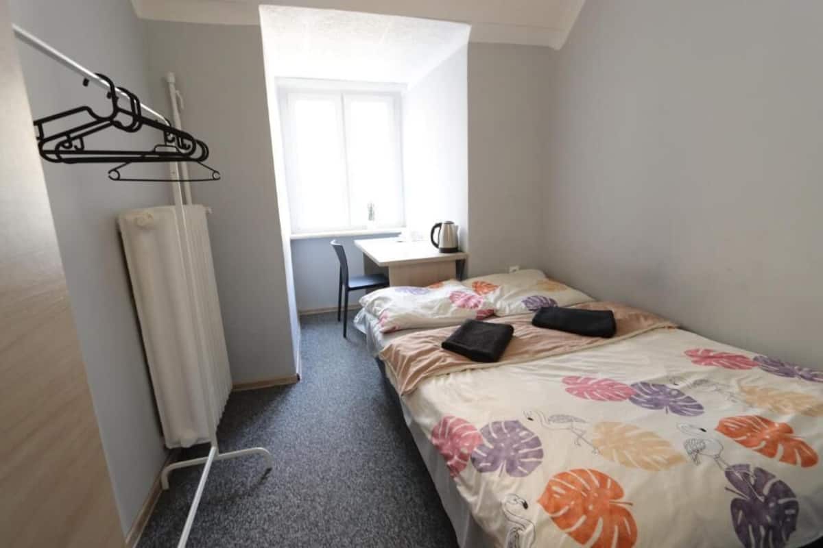 A small, well-lit room with a bed, a desk, and a radiator.