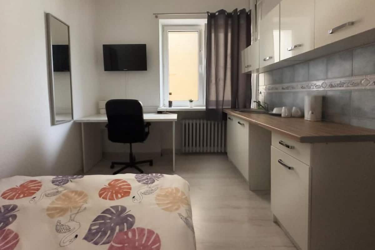 A compact hotel room with a bed, a desk, a chair, a television, a window with curtains, and a kitchenette.