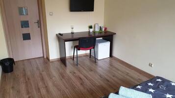 Una habitación con piso de madera, una cama con manta estampada de estrellas, un escritorio con silla, un televisor y un refrigerador pequeño.
