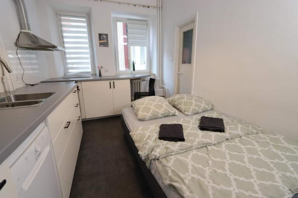 A compact room with a kitchenette, a bed with patterned bedding, and a window with blinds.