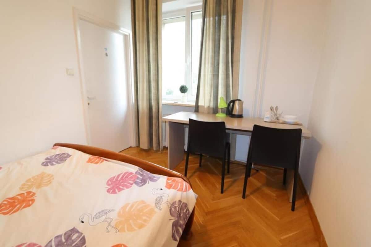 A bedroom with a bed, a desk, two chairs, a window with curtains, and a small table with a kettle and a cup.