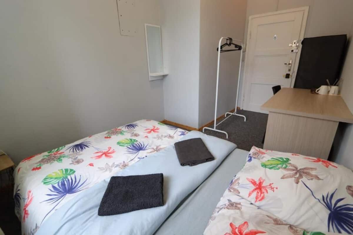 A room with a bed, a bedside table, a desk with a chair, a mirror, and a door.