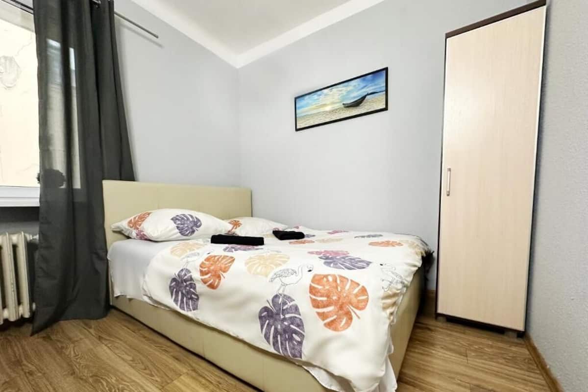 A bedroom with a bed, a radiator, a window with curtains, a painting on the wall, and a door.