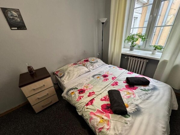 Un dormitorio con cama, mesita de noche, radiador, ventana con plantas y un cartel en la pared.