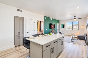 A modern kitchen with a central island, a dining area with chairs, a living room with a sofa, and a view of the outdoors.