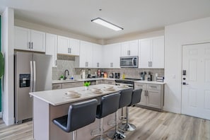 Une cuisine moderne avec des meubles blancs, un îlot central doté de tabourets de bar, un four à micro-ondes encastré et un réfrigérateur en acier inoxydable.