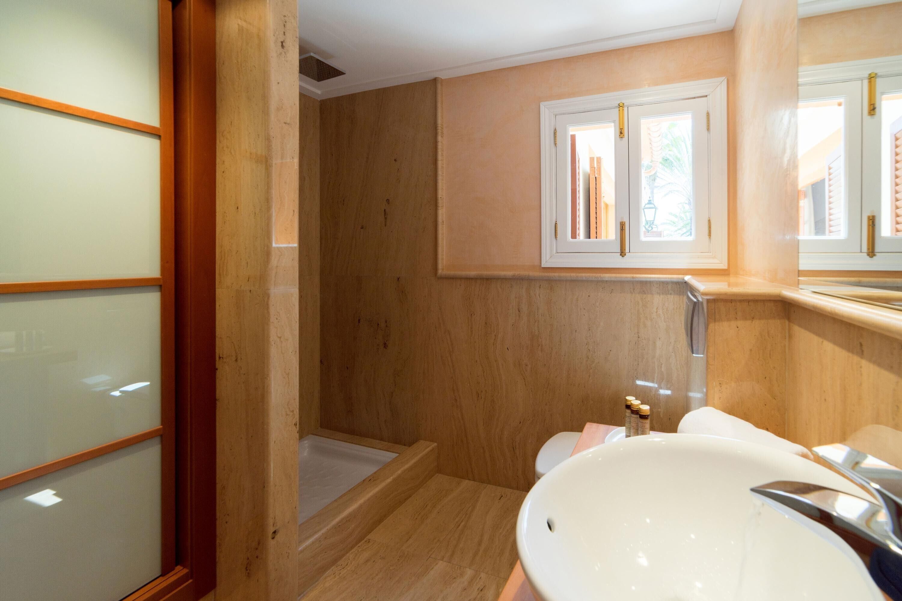 Un baño con piso de madera, un lavamanos blanco, un espejo y una ventana con vista a palmeras.