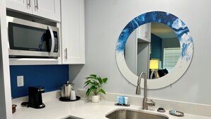 Cocina compacta con microondas, cafetera, lavabo y un espejo redondo con diseño de ola azul.