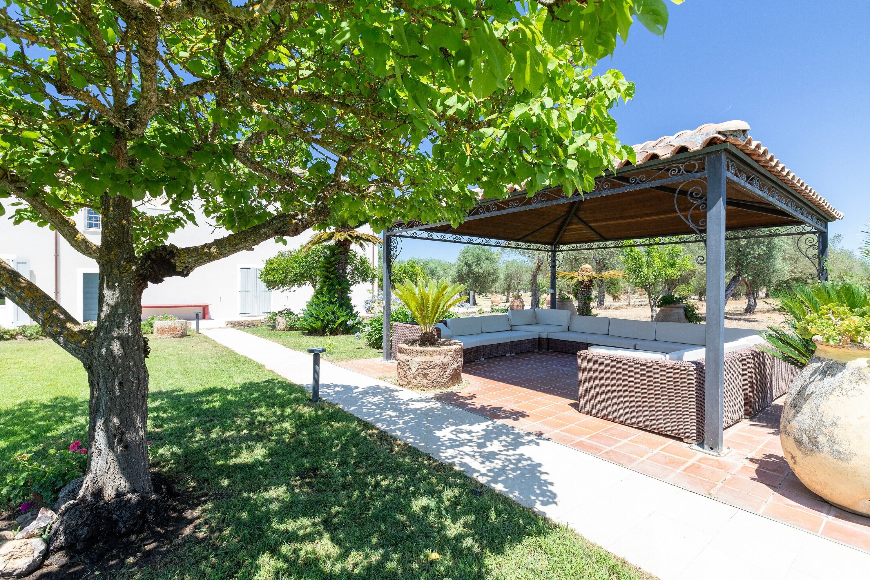 Un espacio de estar al aire libre cubierto con una pérgola, un árbol y un patio con sofá blanco y mobiliario de mimbre.