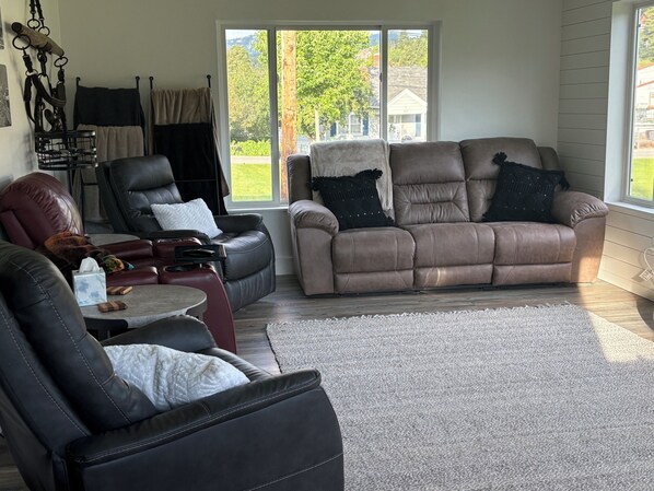 A living room with a brown leather sofa, a black leather armchair, and a red leather chair. There is a small table with a tissue box and a remote control. A window provides a view of a house and greenery outside. - Huge 6-bedroom house in charming Lead w/ plenty of trailer parking, (Lead)