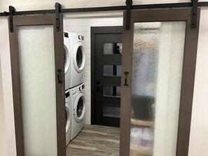 A laundry room with stacked front-loading washers and dryers, a dark door with frosted glass panels, and a wooden floor. - Huge 6-bedroom house in charming Lead w/ plenty of trailer parking, (Lead)