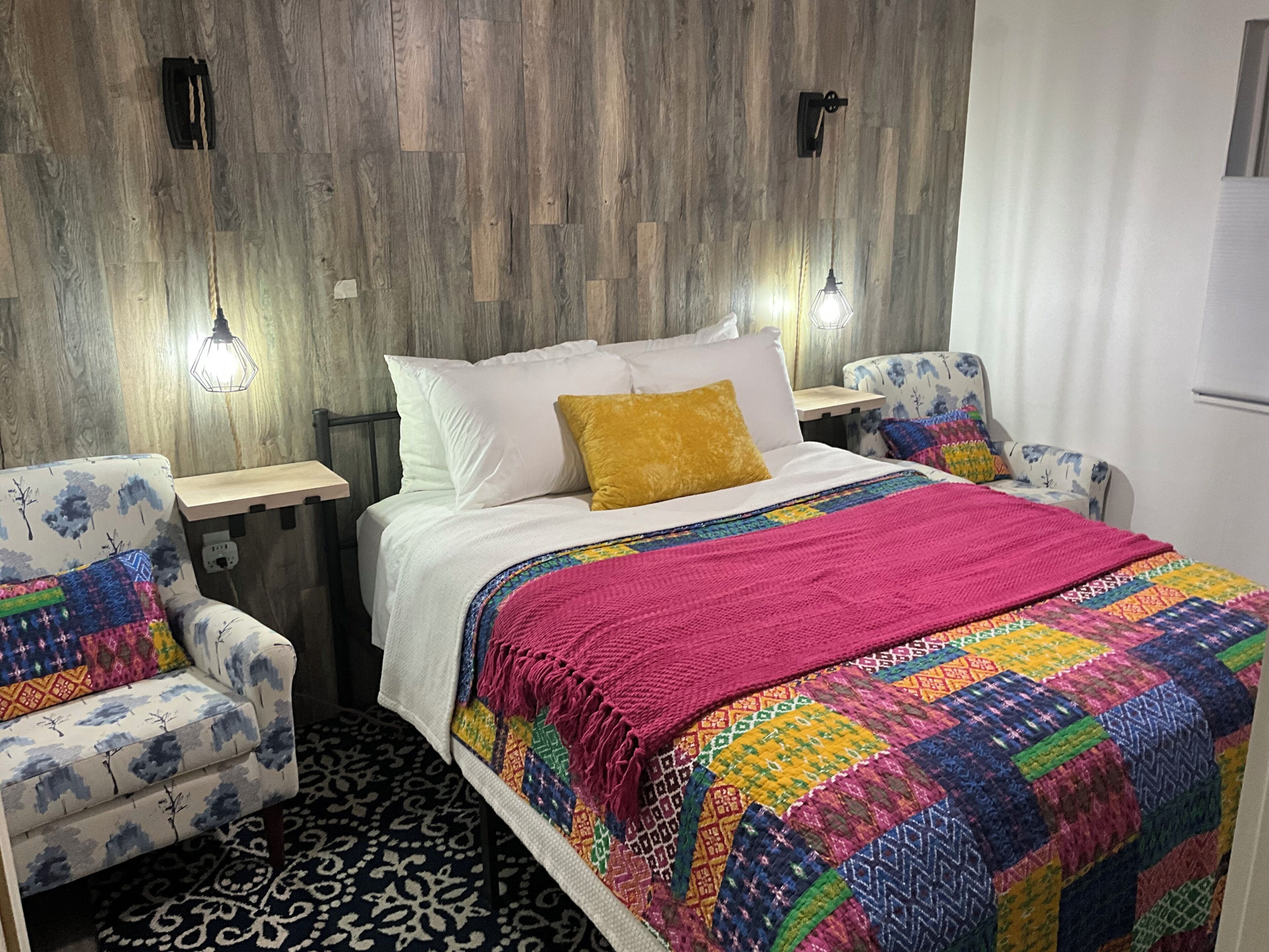 A hotel room with a wooden wall, a bed with a colorful quilt, two patterned armchairs, and two wall-mounted lights.