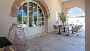 Um alpendre coberto com mesa e cadeiras, uma grande janela em arco e vista para o verde lá fora.