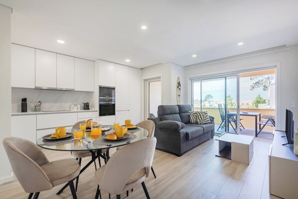 A modern kitchen with white cabinets, a dining table with chairs, a grey sofa, and a TV.