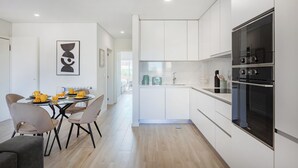 A modern kitchen with white cabinets, a built-in oven, and a dining area with a table and chairs. - The Pine Flat with Pool at Furadouro (Ovar)