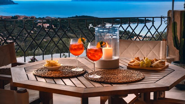 Een houten tafel met twee glazen rode drank, een kaars en een bordje met hapjes op een balkon met uitzicht op de kust.