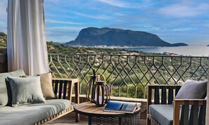 A balcony with a view of a mountain and sea, furnished with a sofa, chairs, and a table with a book and a lantern.