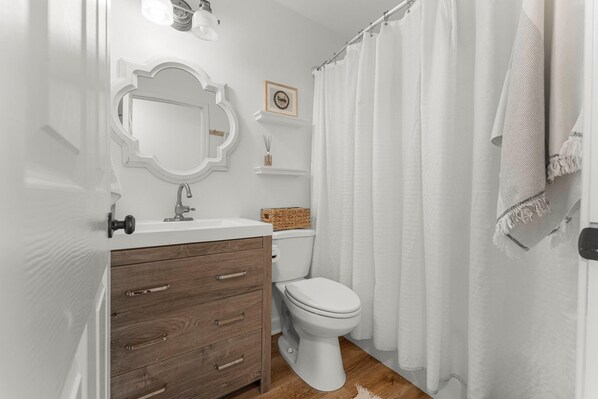 A bathroom with a white toilet, wooden vanity, and a white shower curtain. - Cottage Haven (Santa Rosa Beach)