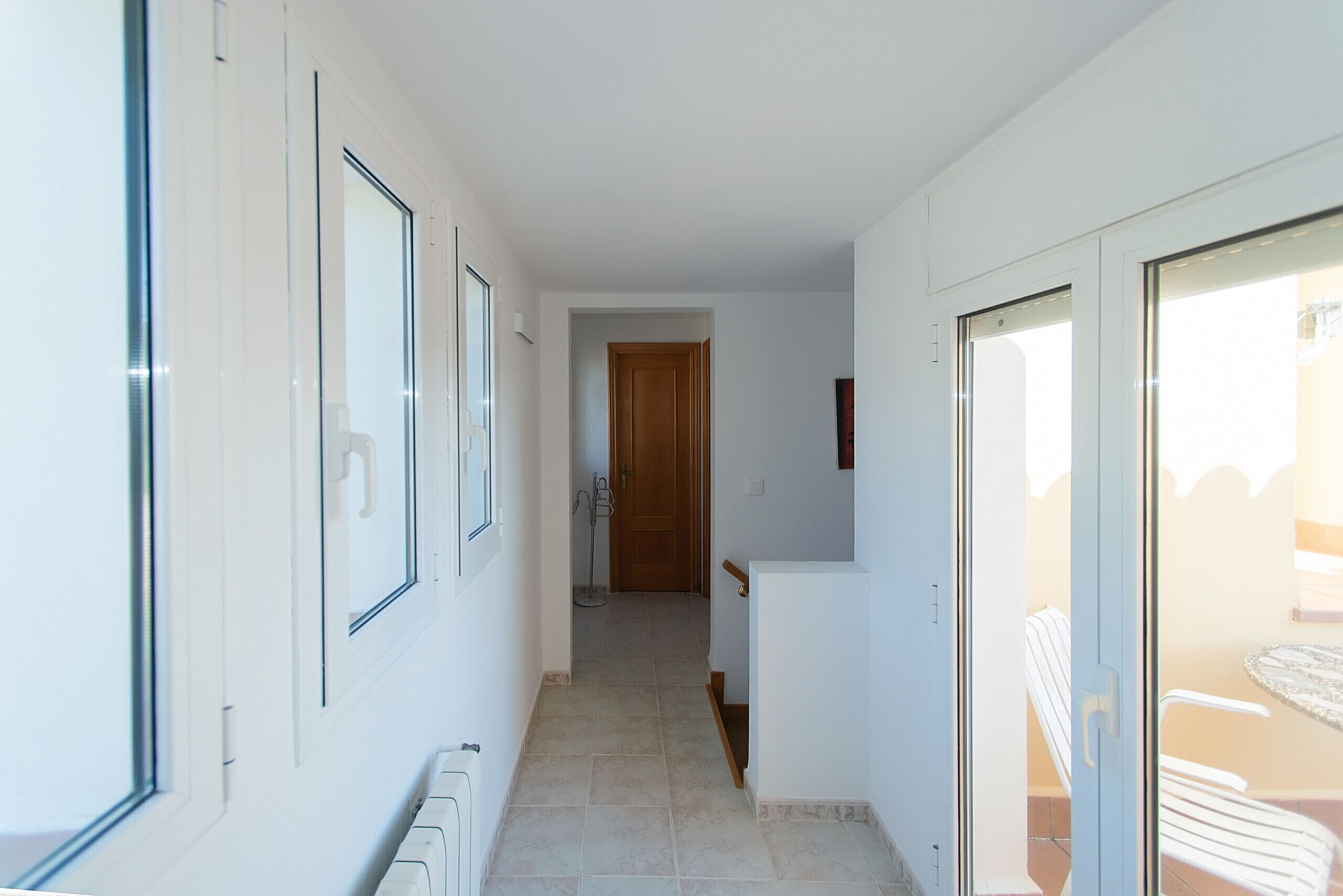 A narrow hallway with white walls, a wooden door at the end, and a glass door leading to an outdoor area with chairs.